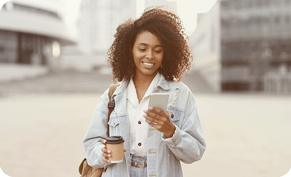 A smiling woman holding a coffee cup checks her phone while walking through a city plaza