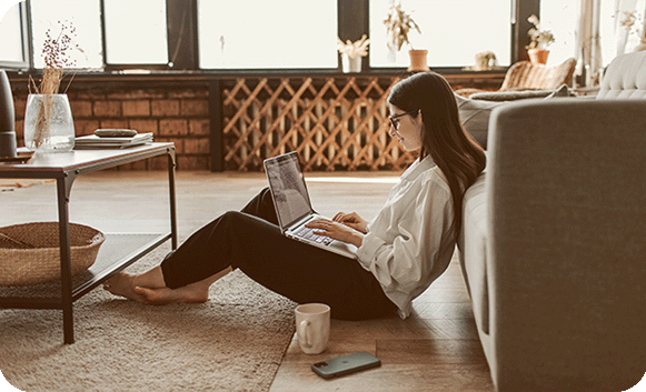 A woman sits on the floor of her living room working on a laptop, with a mug and phone beside her