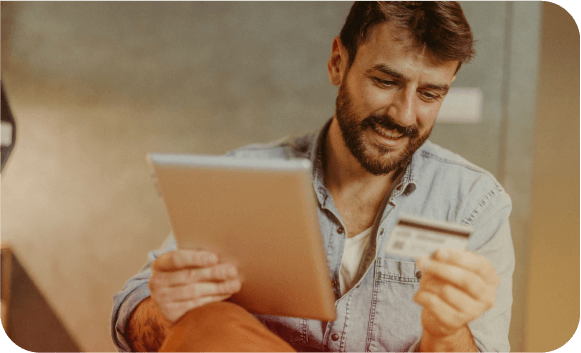 A smiling man holds a tablet in one hand and a payment card in the other while shopping online