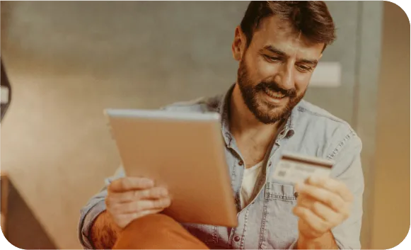 A smiling man holds a tablet in one hand and a payment card in the other while shopping online