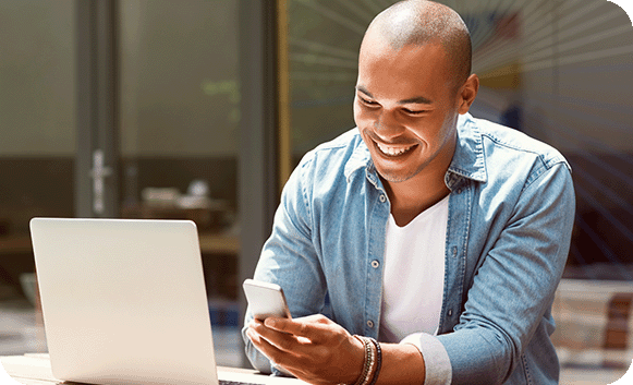 A smiling man in a denim shirt uses his phone at a table beside an open laptop