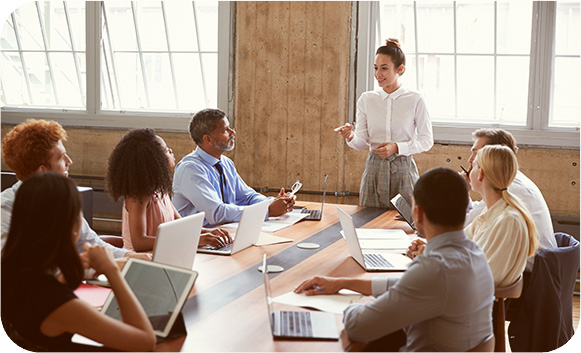 A presenter standing at the head of a meeting table, talking a team with laptops through a plan