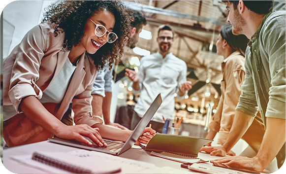 A smiling colleague working on a laptop at a shared desk while teammates collaborate around her